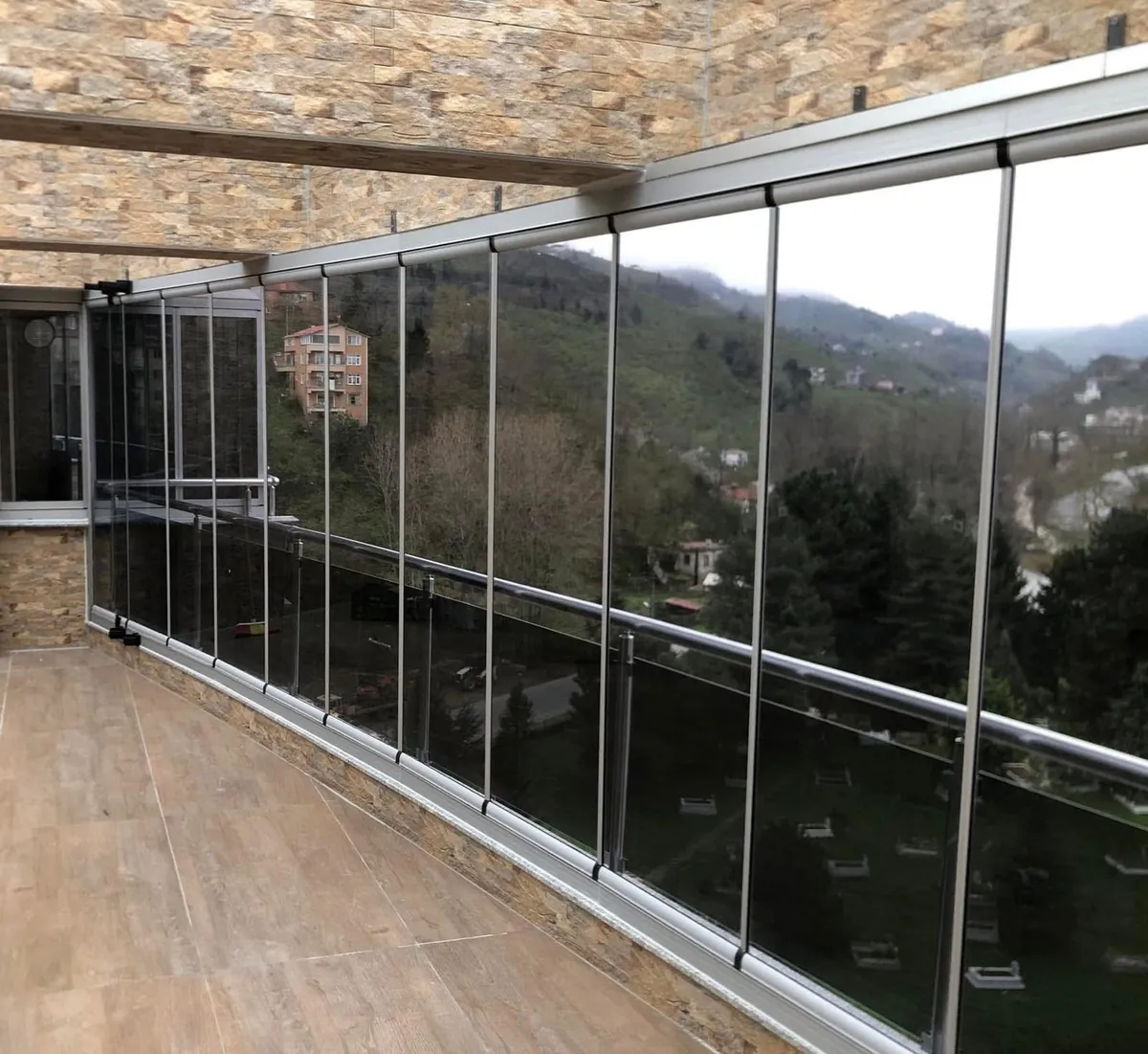Taş kaplı uzun terası baştan başa kapatan mat gri profilli katlanır cam sistem ve arkasındaki cam korkuluk