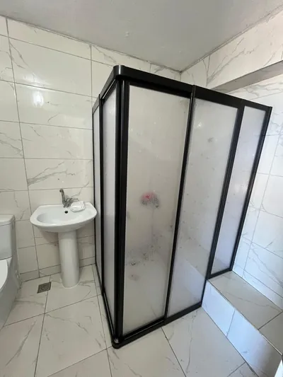 Siyah profilli buzlu camlı duşakabin, beyaz mermer desenli küçük banyoda oturma sekisine bitişik yerleştirilmiş