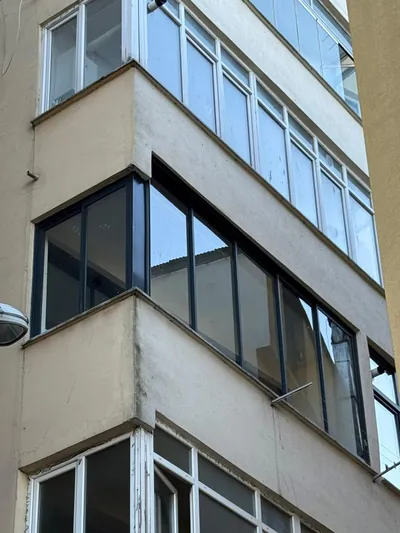 Apartman cephesinde antrasit alüminyum profilli kapalı balkon, üst kattaki eski beyaz kapatmaların arasında köşe dönüyor