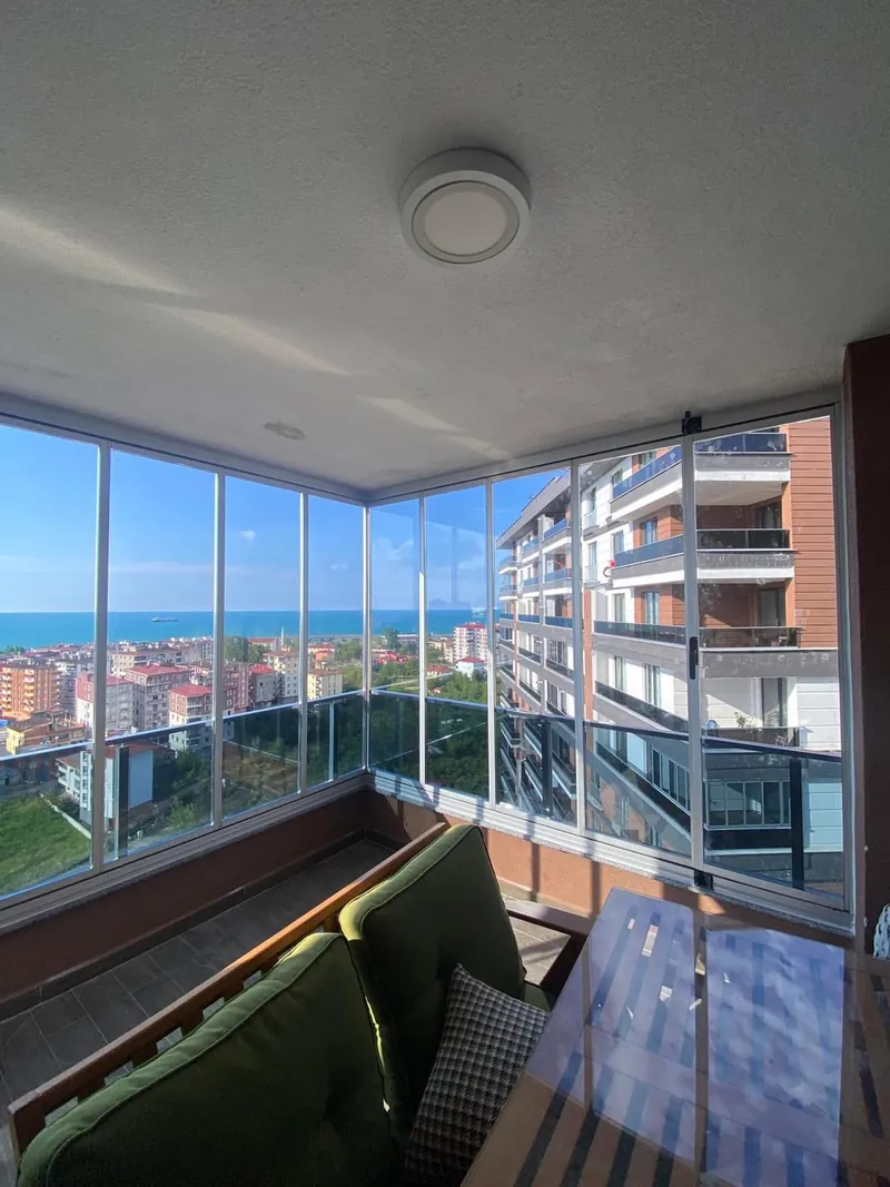 Beyaz alüminyum profilli köşe balkon deniz manzarasına açılıyor, içinde yeşil minderli ahşap kanepe ve cam sehpa