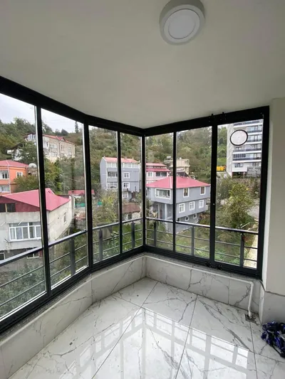 Siyah alüminyum profilli köşe dönüşlü kapalı balkonun içinden yamaç yerleşimine bakış, zeminde mermer desenli seramik