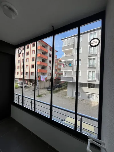 Koyu tonlu iç mekânda siyah profilli çoklu kanatlı sürme cam sistem, camların ardında sokak ve komşu apartmanlar